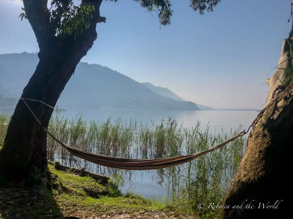 A serene lakeside scene with a hammock tied between two trees, overlooking a calm Lake Mutanda with hills in the soft haze of the background. Plan a day in your Uganda itinerary to rest at Mutanda Lake Resort.