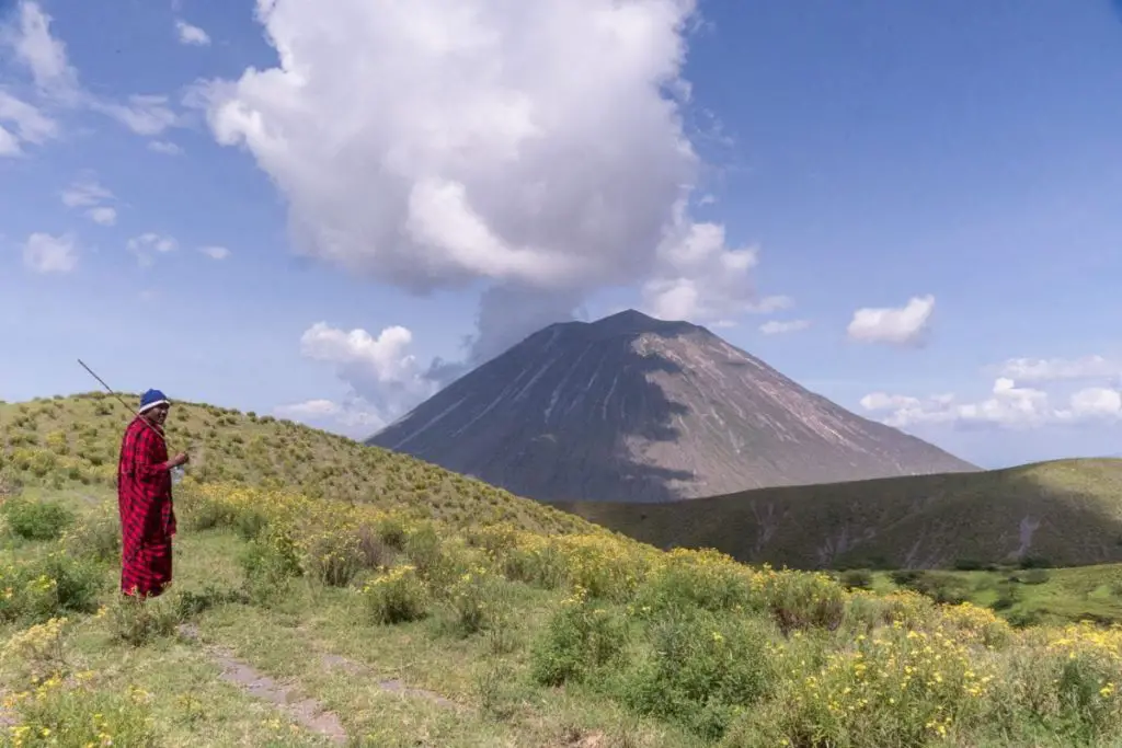 A Maasai man in traditional red clothing and a blue hat stands on a grassy hillside, looking towards a towering volcano against a blue sky with clouds.