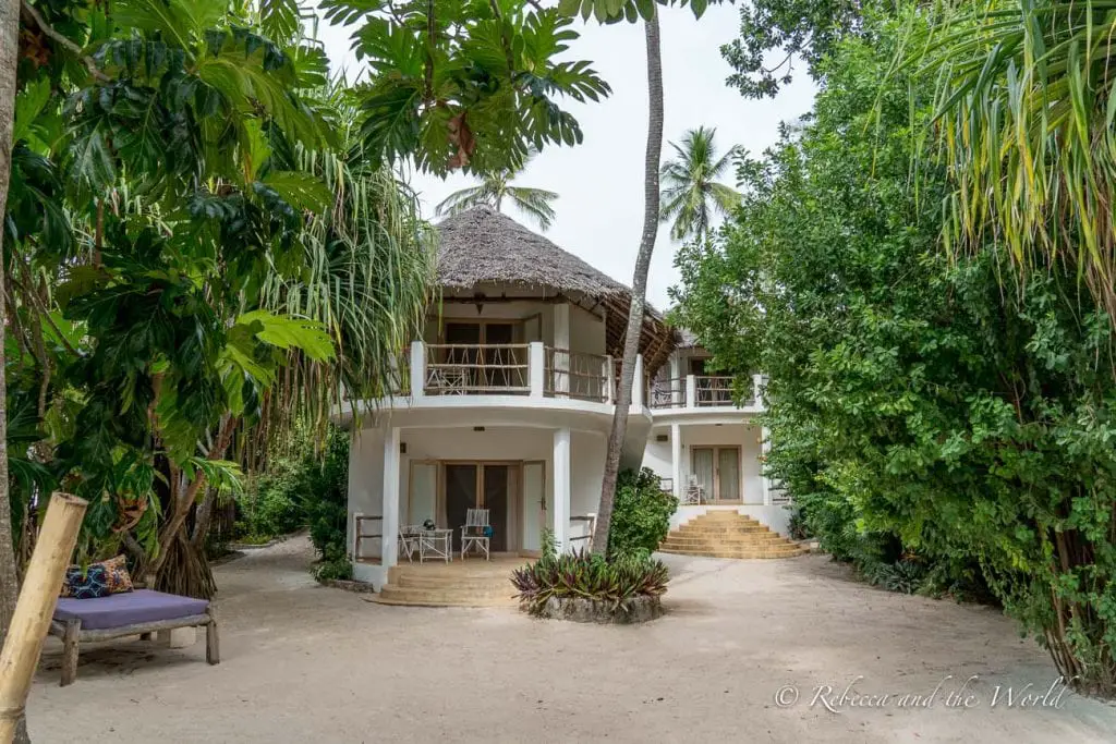 A two-story white building with a thatched roof, surrounded by lush tropical vegetation, with chairs on the porch. Sharazad Boutique Hotel in Jambiani is a beautiful place to stay in Zanzibar.