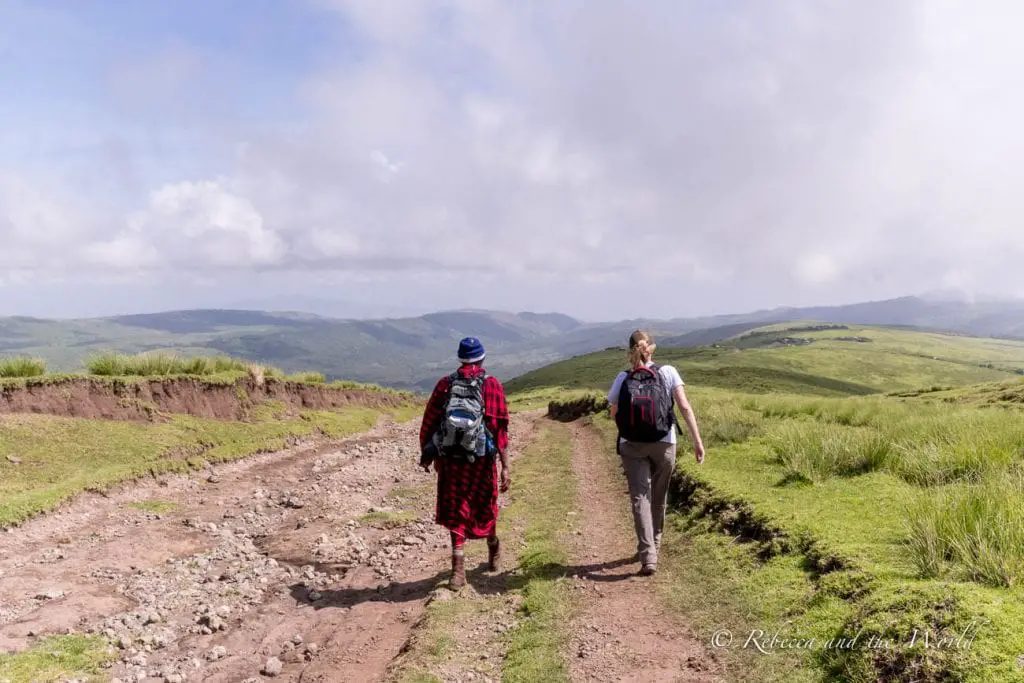 Two people walking on a dirt path in a hilly landscape. One is a Maasai man wearing a red garment and a backpack, the other in casual hiking attire. One of the most incredible things to do in Tanzania is a 3-day hike from Ngorongoro to Lake Natron.