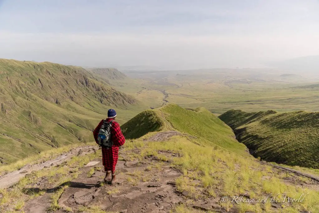 A Maasai man in traditional red clothing and a backpack walks along a narrow dirt path on a green ridgeline with expansive views of the valley below. One of the best things to do in Tanzania is a hike from Ngorongoro to Lake Natron.