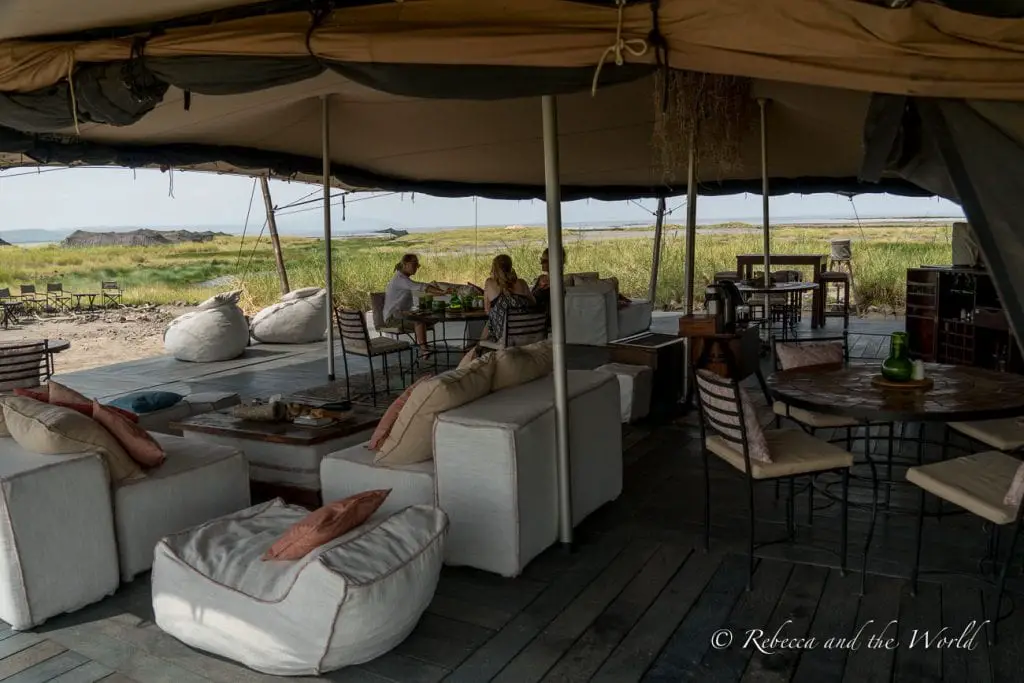 A comfortable outdoor lounge area under a large tent with white couches and cushions, with a view of the open plains in the background. While Lake Natron Camp is remote, they have created a unique experience that is also eco friendly.