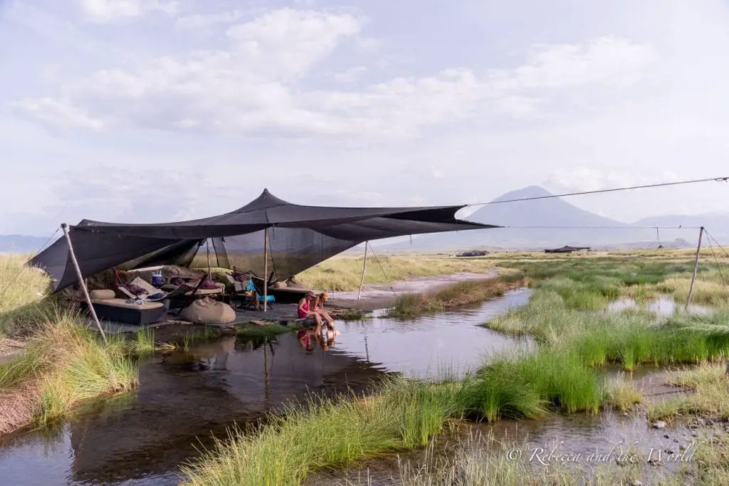 An open tented structure with lounge chairs, set by a small stream in a grassy landscape, with hills in the distance. While Lake Natron Camp is remote, they have created a unique experience that is also eco friendly.