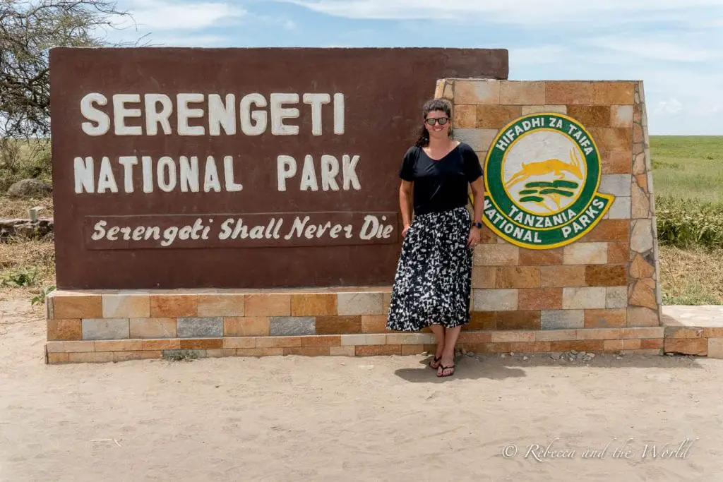 A woman (the author of this article) stands next to a sign reading "SERENGETI NATIONAL PARK" with the slogan "Serengeti Shall Never Die" and the emblem of Tanzania's national parks. One of the best places to visit in Tanzania is the Serengeti.