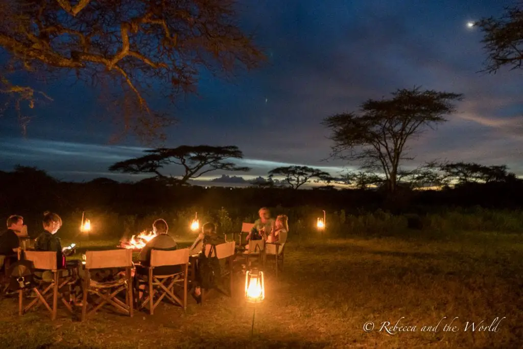 A group of people dining outdoors at night, illuminated by lanterns, under a tree and a twilight sky. One of the best places to stay in Ndutu in Tanzania is Lemala Ndutu, a mobile tented camp.