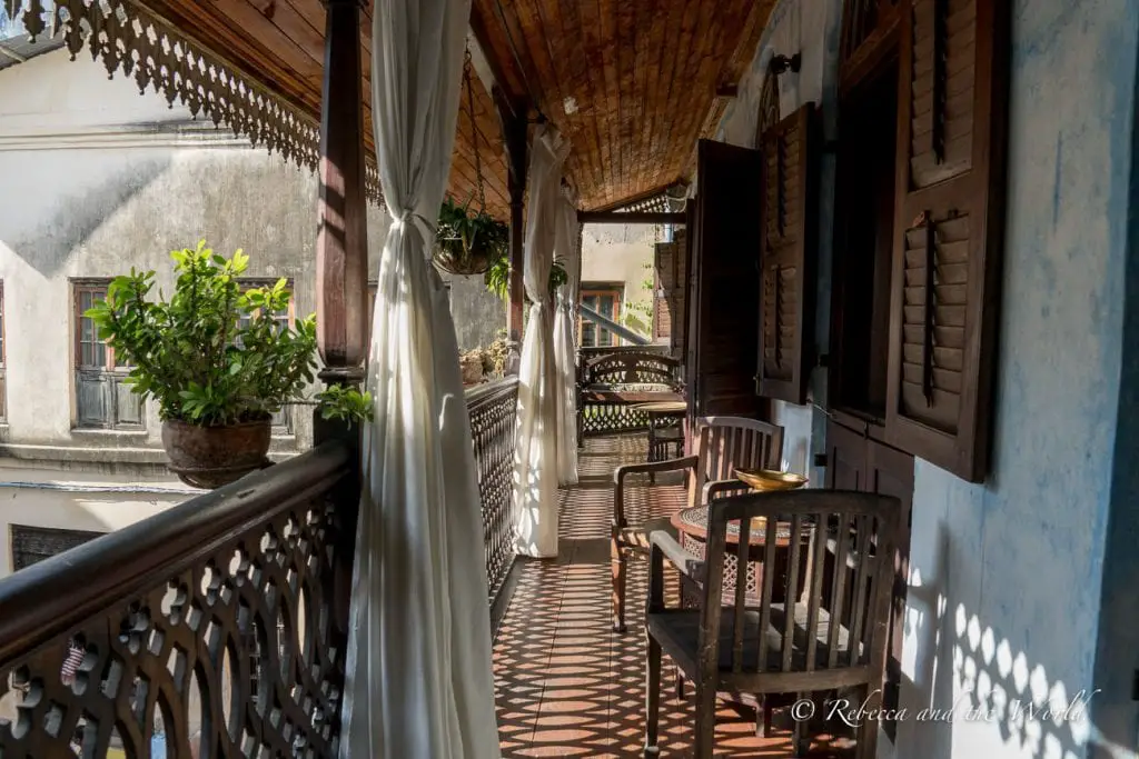 A shaded veranda with wooden chairs and hanging white curtains overlooking an urban landscape. The Emerson Spice Hotel is one of the best places to stay in Stone Town in Zanzibar.
