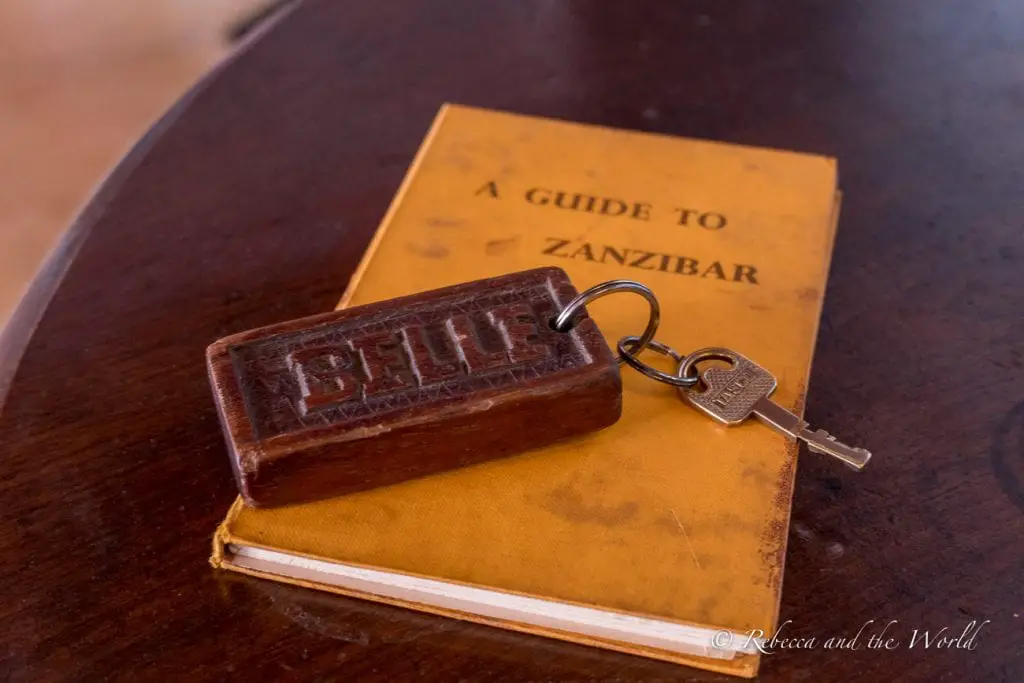 A vintage-style room key with an embossed wooden tag reading "BELLE" lies atop a weathered yellow book titled "A Guide to Zanzibar" on a wooden surface. The Emerson Spice Hotel is one of the best places to stay in Stone Town in Zanzibar.