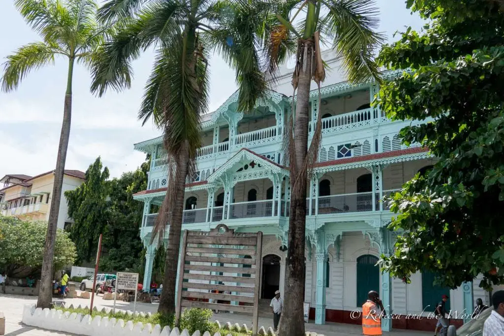 A large, ornate, mint-green building with intricate woodwork and balconies, framed by tall palm trees under a blue sky. One of the best things to do in Stone Town in Zanzibar is a walking tour of the city.