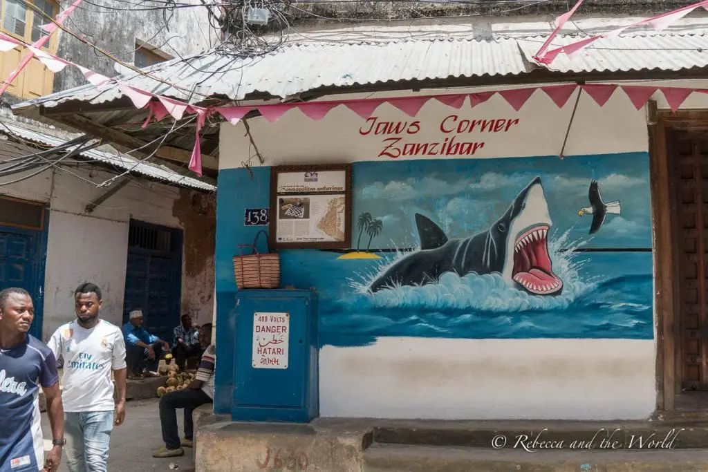 A mural of a large shark, titled "Jaws Corner Zanzibar," on the wall of a building with people walking by in front. One of the best things to do in Stone Town in Zanzibar is a walking tour of the city.