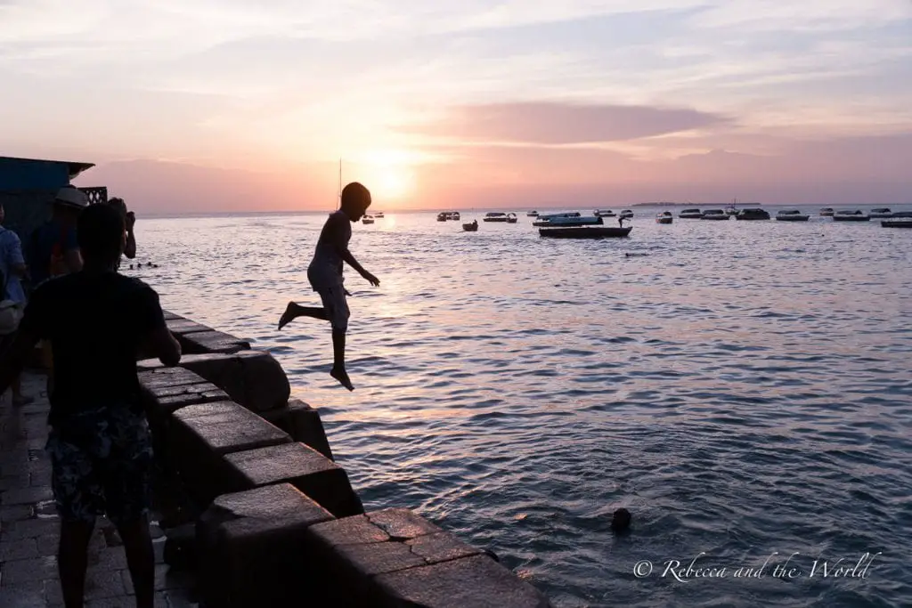 A silhouette of a person leaping off a seaside wall into the ocean at sunset, with boats in the distance. At sunset each evening, Forodhani Gardens are filled with young boys jumping off the walls into the water below.