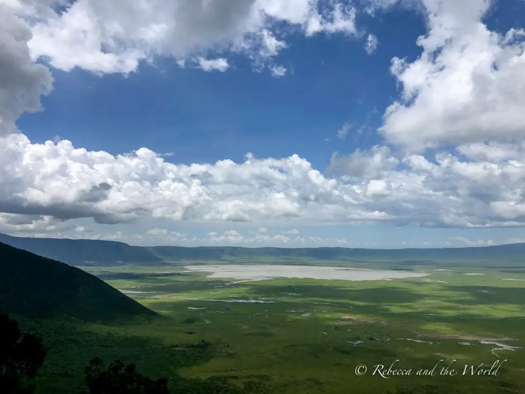 A panoramic view of the vast, green Ngorongoro Crater with a lake in the distance. The sky above is partially covered by fluffy clouds. The Ngorongoro Crater in Tanzania is a huge caldera caused by a volcanic eruption more than 2 million years ago.