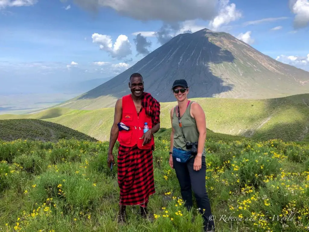 Two people, a Maasai man in traditional red clothing and the other a woman (the author of this article) in casual outdoor attire, stand smiling in a field of yellow wildflowers with a volcanic mountain in the background. Walking past Ol Doinyo Lengai, Tanzania's only active volcano, is a highlight of the hike from Ngorongoro to Lake Natron.