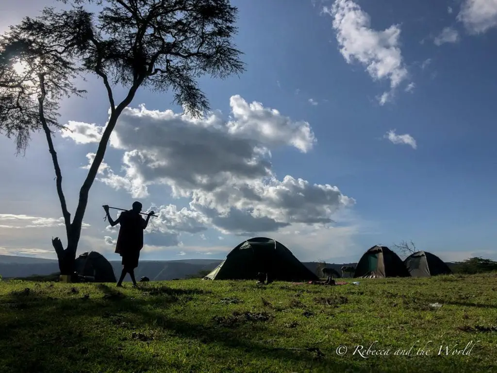 A silhouette of a person standing next to a tree, holding a long stick, with several tents set up in the background under a sky with scattered clouds. Acacia Forest Camp is the camping spot for night 2 of the hike from Ngorongoro to Lake Natron.