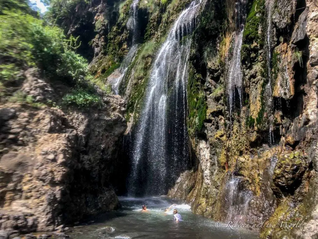 Several individuals swimming at the base of a cascading waterfall surrounded by rocky cliffs and greenery. Lake Natron Camp can arrange a hike to nearby waterfalls.