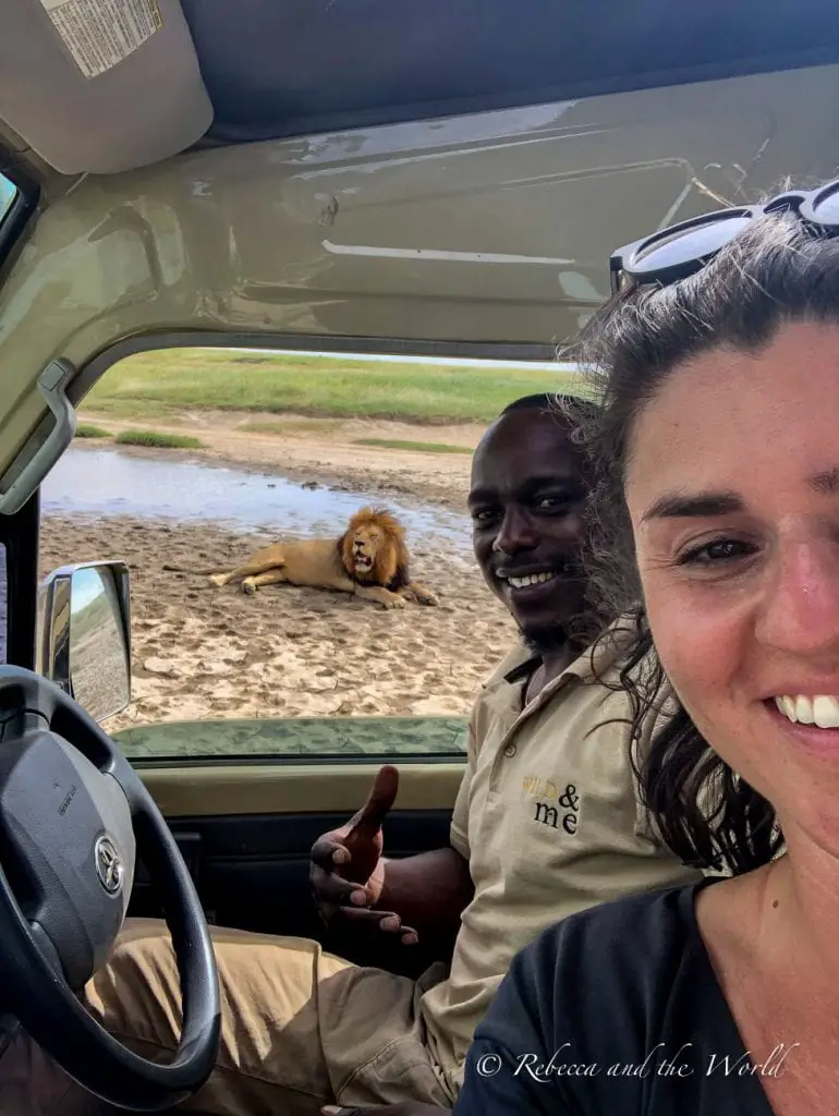 Two people are inside a vehicle; a woman (the author of the article) on the right taking a selfie and a man on the left doing a thumbs-up. Outside the vehicle, a lion lies on the ground. Lion selfie with my guide, Baraka.