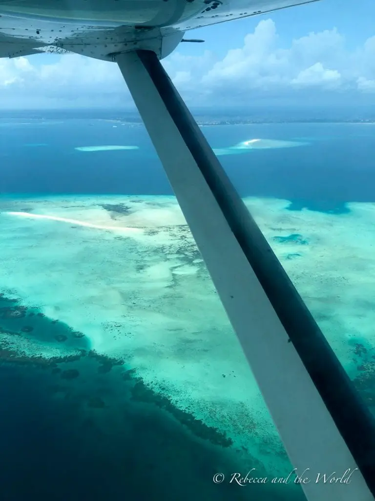 The image shows a view from the window of a small airplane as it comes into Zanzibar, looking down over a turquoise and blue sea with varying shades indicating different depths. Patches of coral reefs are visible beneath the surface of the water, and the wing of the plane is prominently featured in the top left of the frame, creating a sense of flight and perspective over the stunning aquatic landscape. No Tanzania itinerary is complete without a visit to Zanzibar.