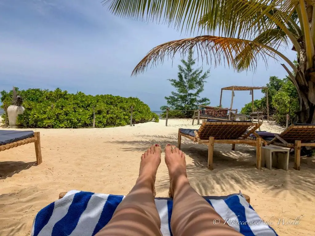 The viewpoint of a person (the author of this article) lounging on a beach chair on a sandy beach with a view of sun loungers, palm trees, and a clear sky. One of the best things to do in Zanzibar is relax on the beach.