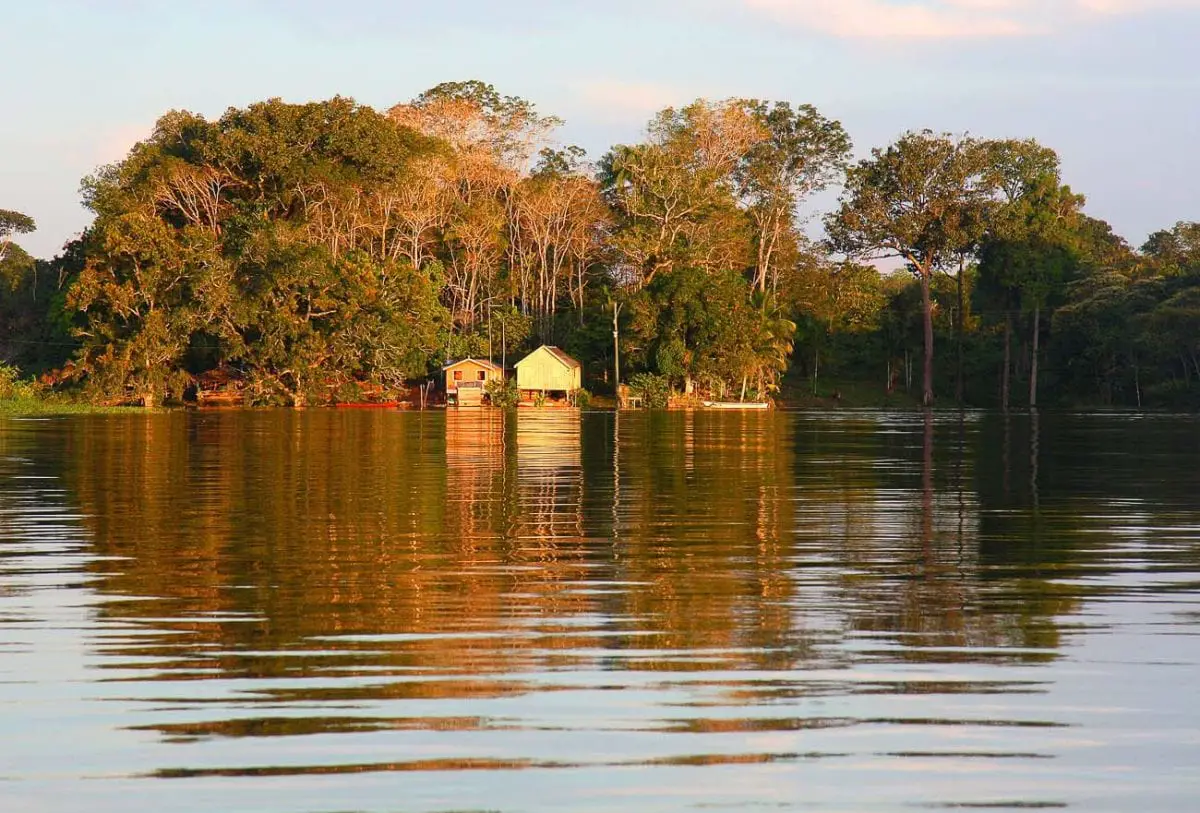 A serene riverside setting in the Amazon, with a small stilt house surrounded by dense jungle reflecting in the calm waters at sunset. One of the places that has long been on my bucket list is the Amazon.