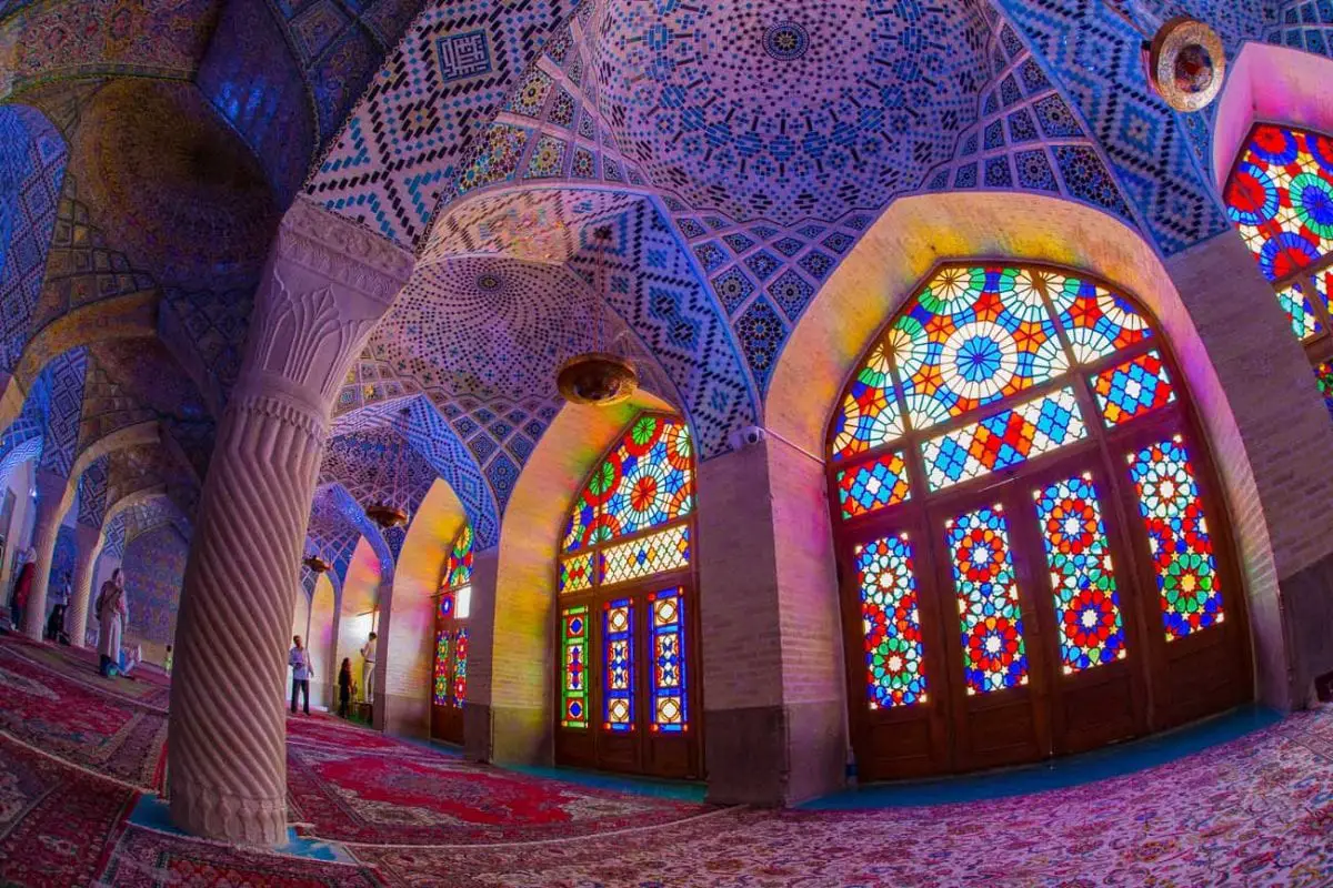 The interior of the Nasir al-Mulk Mosque in Iran, featuring a stunning display of intricate Islamic architecture, colorful mosaics, and stained glass that casts vibrant light patterns onto the carpeted floor. When it's safe to travel, visiting Iran is on my bucket list.