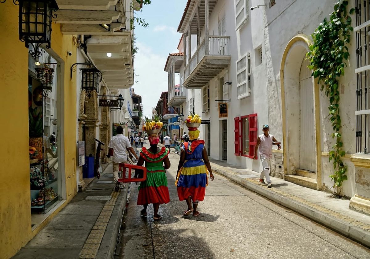 Two women dressed in traditional, colorful Colombian attire with fruit baskets on their heads, walking away from the viewer down a colonial street in Cartagena, evoking the country's vibrant culture. One of my bucket list countries is Colombia.