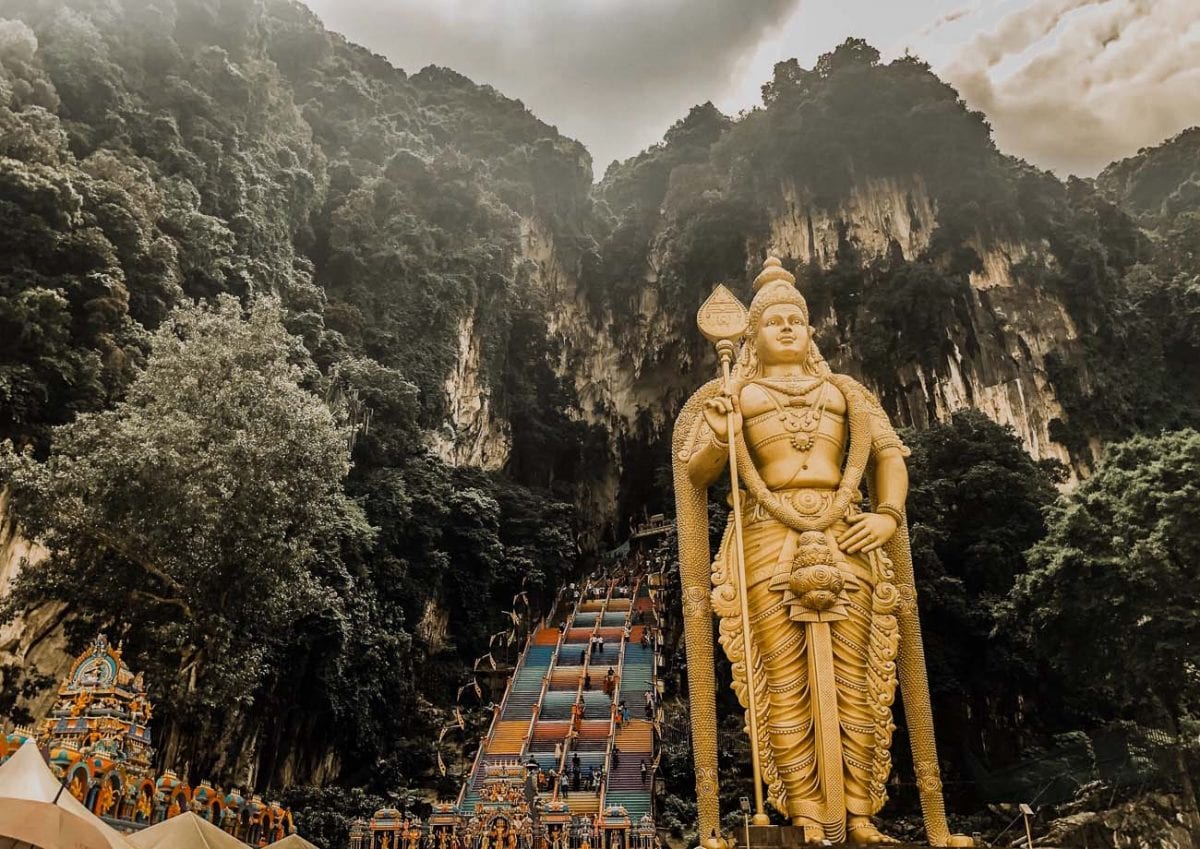 The towering golden statue of Lord Murugan at the entrance of the Batu Caves in Malaysia, with a colorful flight of stairs leading up into the limestone hill, surrounded by lush greenery. Malaysia is known for its culture, food and beaches.