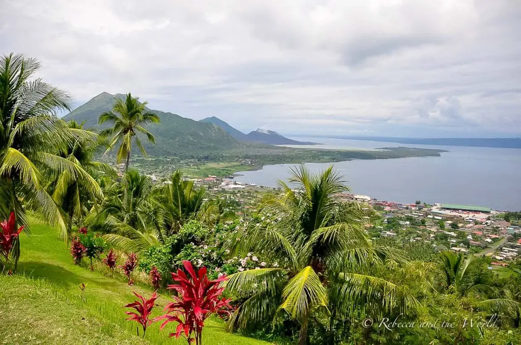 A lush tropical landscape viewed from a hilltop, showing a dense spread of palm trees and foliage, with a small coastal town by the water's edge and mountains in the background. Papua New Guinea is a country full of bucket list adventures.