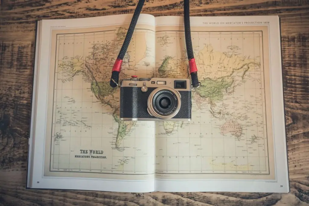 A vintage-style camera with a black strap rests atop an open atlas displaying a Mercator projection map of the world. The setting conveys a theme of travel and exploration.