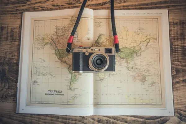 A vintage-style camera with a black strap rests atop an open atlas displaying a Mercator projection map of the world. The setting conveys a theme of travel and exploration.