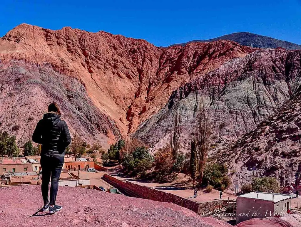 A person - the author of this article - in a black jacket and jeans stands overlooking a valley with striking multicolored mountain formations, showcasing the vivid and natural geological beauty. This is the town of Purmamarca in northern Argentina.