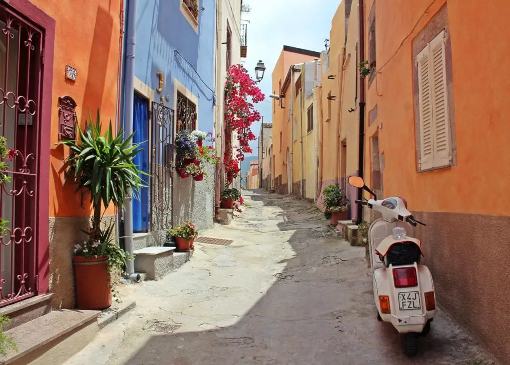 Narrow cobblestone street in a quaint European village, with colourful buildings and a white scooter parked to one side. The food, beaches and small villages are just a few reasons why I want to visit Italy.