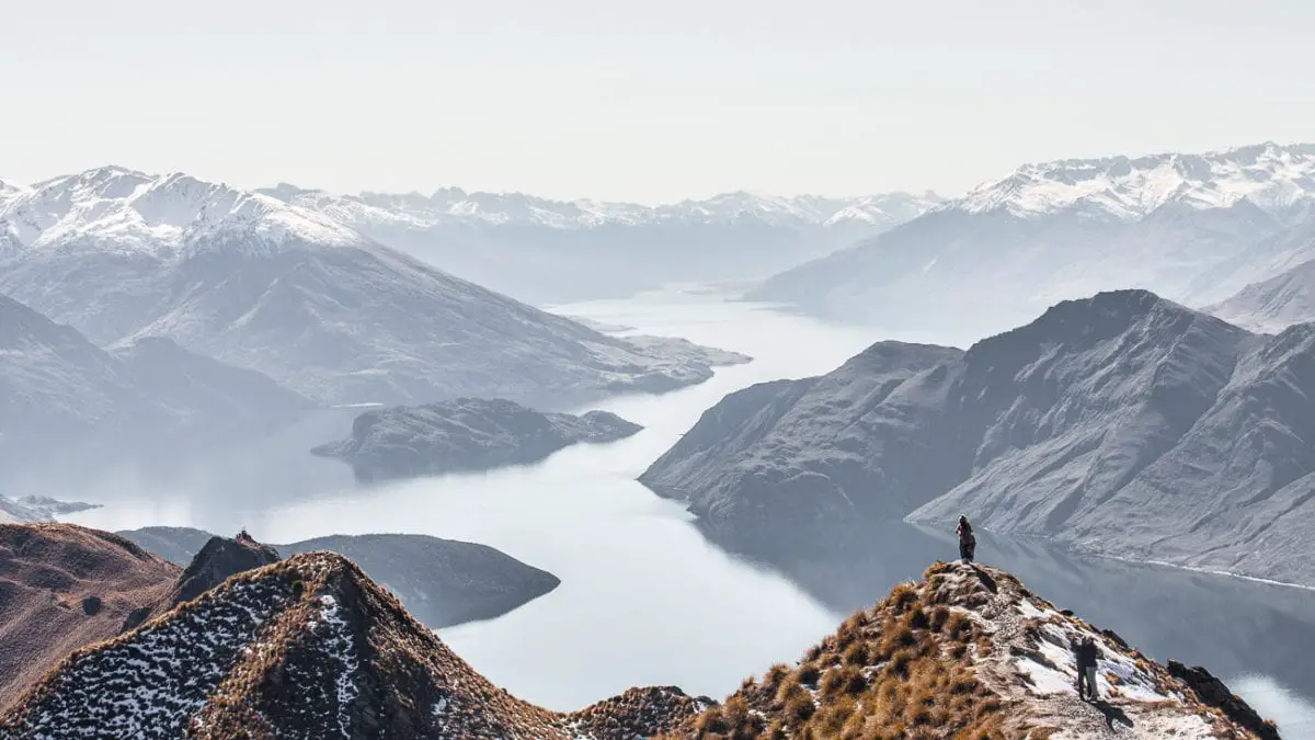 A hiker stands on a mountain ridge overlooking a vast, mist-covered lake surrounded by rugged mountains, emphasizing the scale and grandeur of the natural landscape. New Zealand is one of the most beautiful countries in the world and worthy of adding to any travel bucket list.