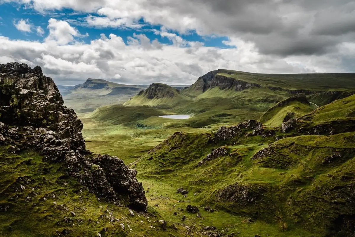 A scenic view of the Isle of Skye, Scotland, with rugged terrain, green valleys, and a small loch nestled amongst the rolling hills under a dynamic sky. The rugged beauty and a family connection are reasons why I'd like to visit Scotland.