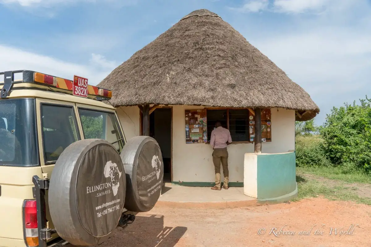 A safari guide standing at a small service window of a thatched-roof building, with a safari vehicle parked nearby. One of the reasons why you should hire a private guide for a safari in East Africa is because they'll do all the organising - here's Shaun arranging yet another national park entrance permit.