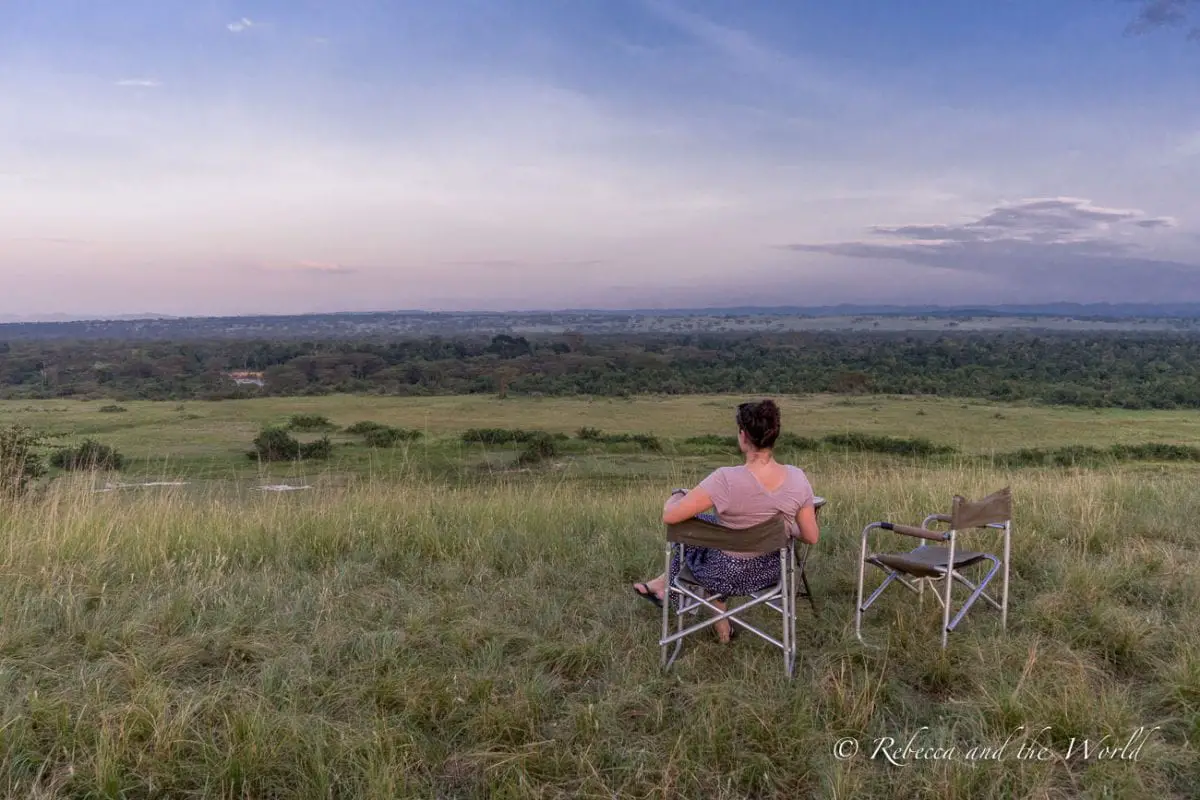 A woman (the author of this article) sitting in a folding chair on a grassy hill overlooking a vast savanna at sunset, representing peaceful moments experienced during a safari. Enjoying a sundowner in Uganda.
