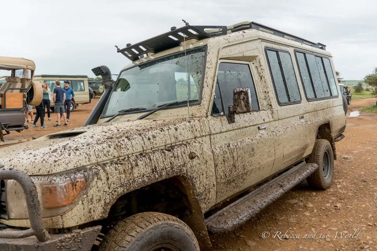 A mud-splattered safari vehicle parked on a dirt road with tourists gathered around, illustrating the adventurous and often rugged conditions of safari travel. Our muddy car after driving through muddy roads in Ndutu in Tanzania.