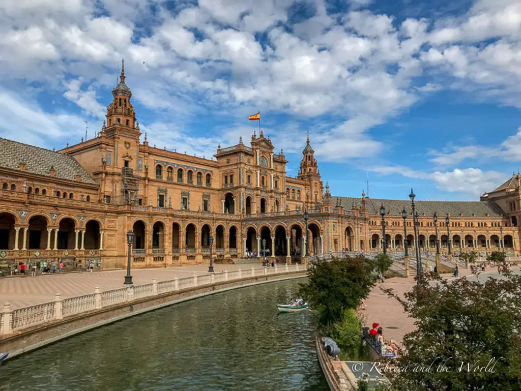 The expansive Plaza de España in Seville, Spain, with its ornate architecture, a canal with a small boat, and visitors walking along the semi-circular building's arcade.