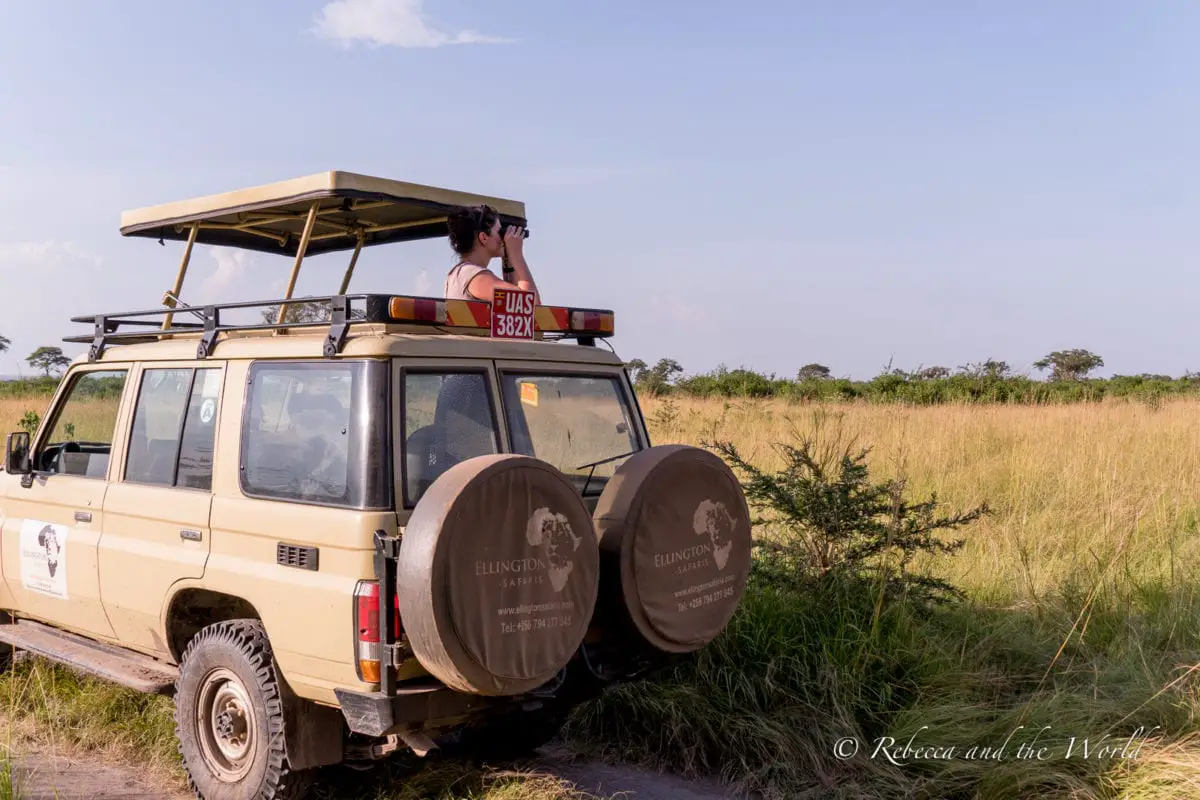 You'll be spending a lot of time in a vehicle while on safari in Africa, so if you're wondering what to wear on safari, my answer is lots of comfy clothes! A woman stands in an Ellington Safaris safari vehicle with binoculars, looking at something in the distance.