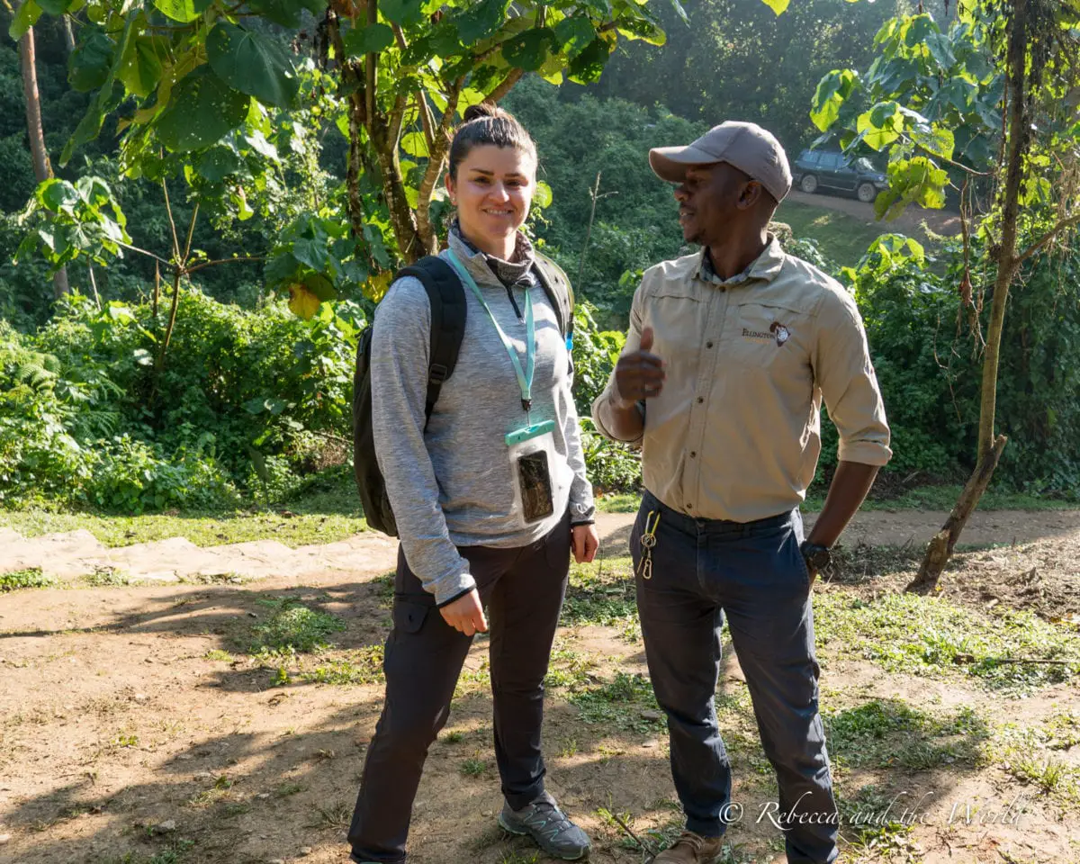 A woman in black pants and a grey sweater stands next to a man in black pants and a khaki shirt. She is looking at the camera and he is looking at her. She is about to start a gorilla trek in Uganda.