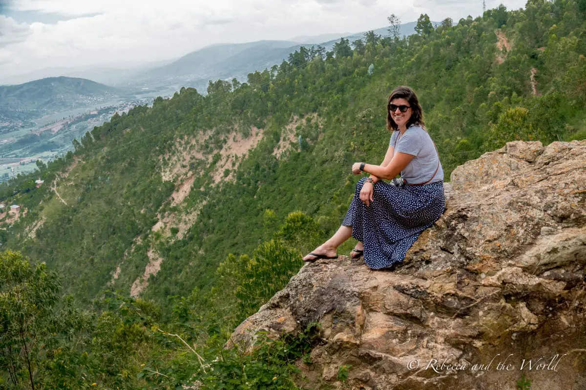 A woman sits on a rocky hill in Kigali, Rwanda, with a view of hills covered in trees. She is wearing a grey top, sunglasses and blue skirt with white spots, and is smiling at the camera.