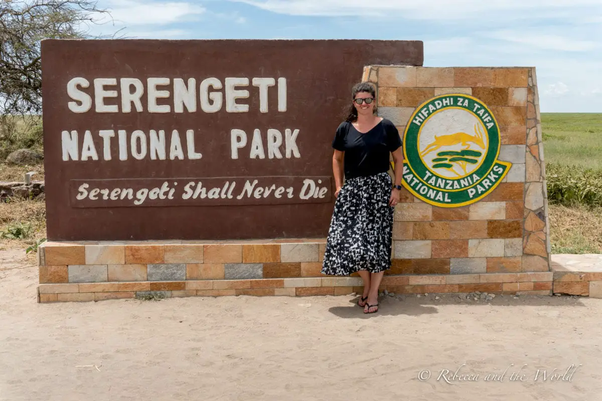 A woman in a black top and black and white skirt stands in front of the sign to the entrance to Serengeti National Park, she is smiling and showing an example of what to wear on safari.