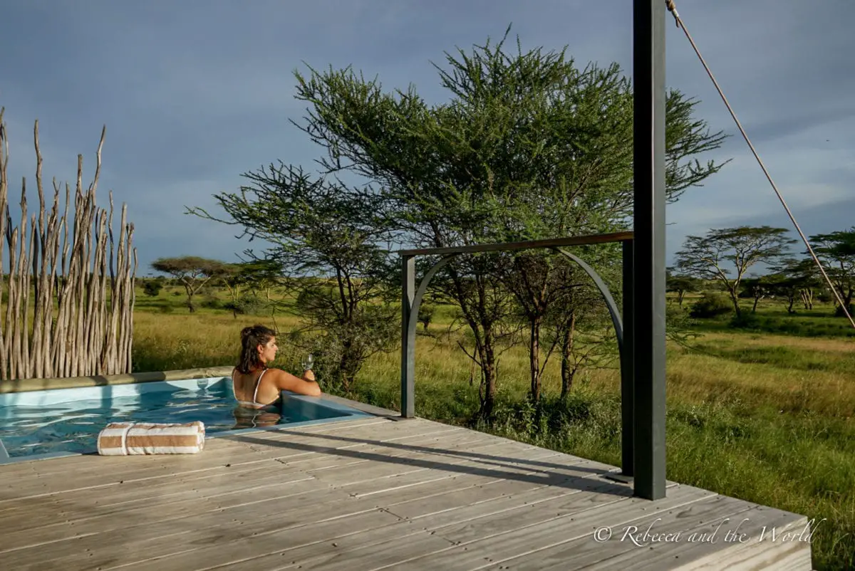 A woman stands in a small plunge pool set into a deck. She holds a glass of wine and there is a brown and white towel on the deck next to the pool. She is looking out to the Tanzanian landscape which is filled with grasses and trees. A bikini is a must on an East Africa packing list to enjoy the beach or swimming pools.