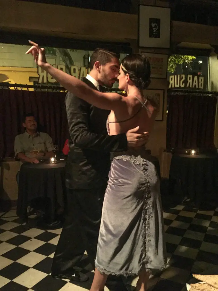 A couple passionately dancing the tango, the man in a suit and the woman in a satin dress, in a dimly lit Buenos Aires dance hall with spectators in the background. See a tango show in Buenos Aires at Bar Sur.