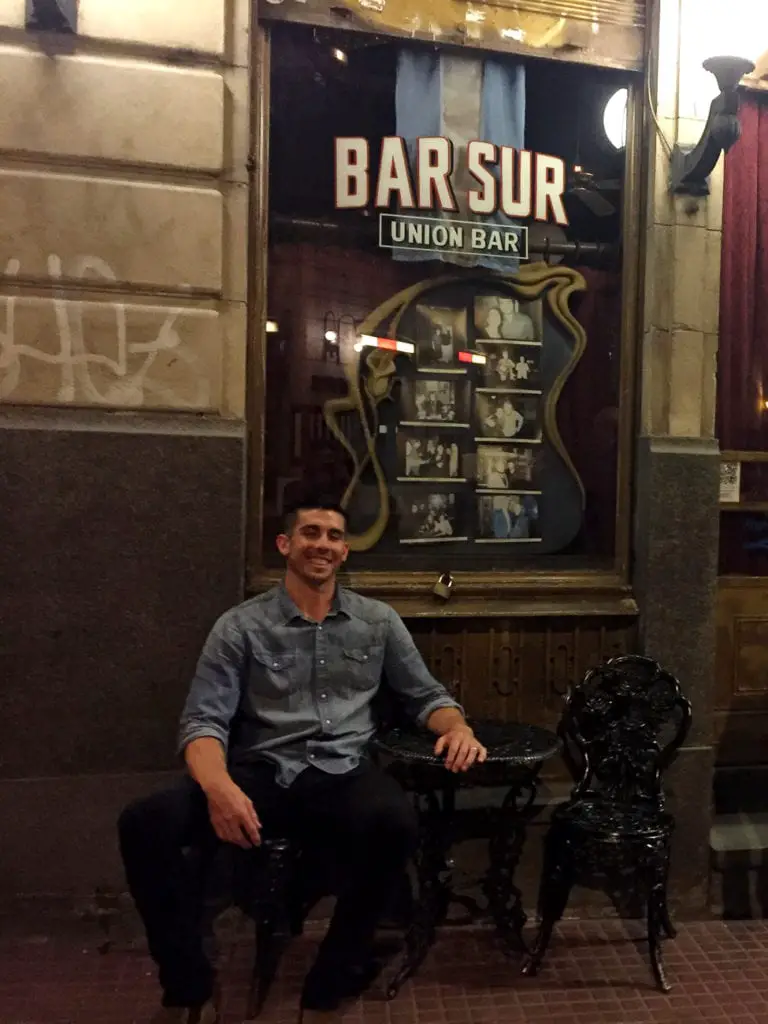 A relaxed man - the author's husband - sitting outside the entrance of the Bar Sur in Buenos Aires, a traditional tango bar, with its vintage sign overhead. See a tango show in Buenos Aires at Bar Sur.