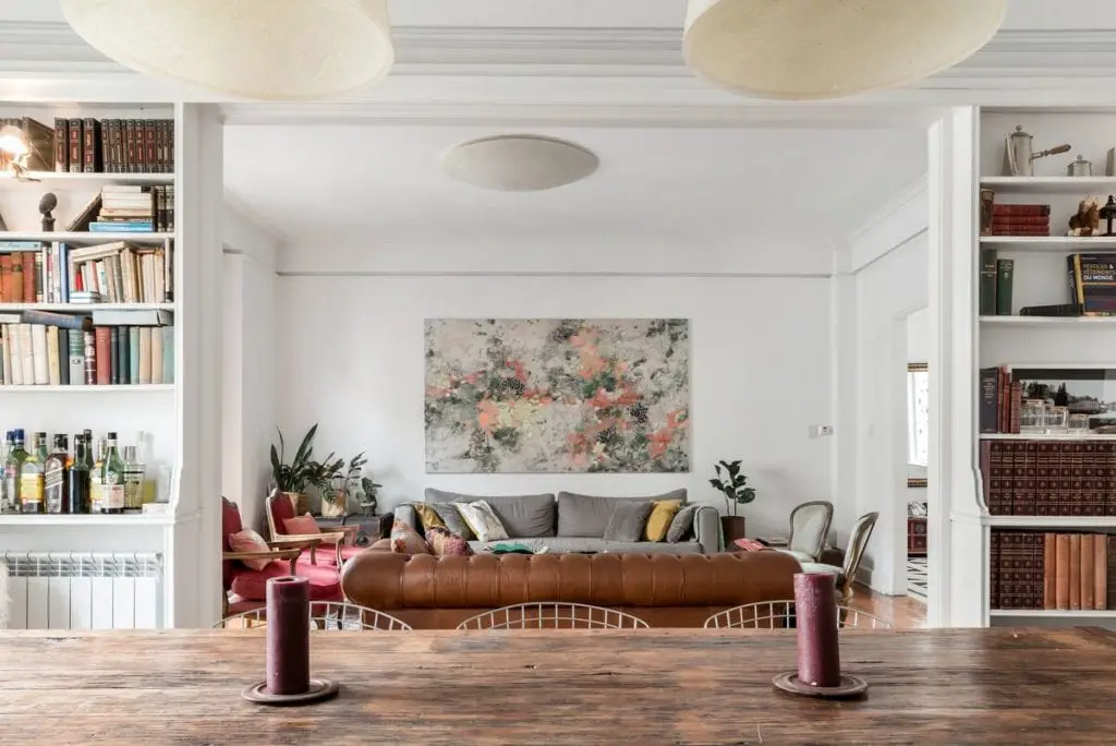 A cozy, well-lit living room with a large brown leather sofa, bookshelves filled with books, and a large abstract painting on the wall.