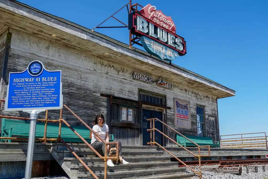 A woman - the author of this article - and a dog seated on the steps of the Highway 61 Blues Visitor Center, with a rustic building backdrop featuring a large 'Gateway to the Blues' neon sign. The entrance to the Gateway to the Blues museum in Tunica, Mississippi.