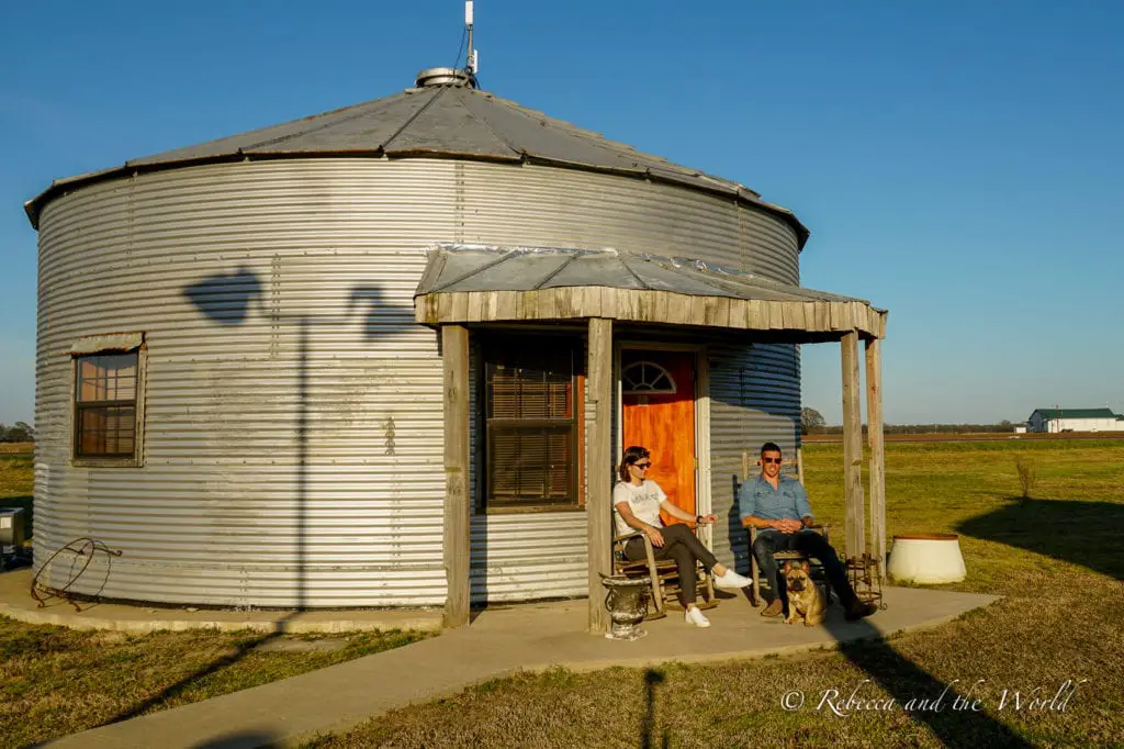 A corrugated metal grain silo converted into a dwelling, with two people - the author and her husband - and a dog sitting on chairs outside, enjoying the expansive flat farmland surrounding them. Stay at the Shack Up Inn in Clarksdale Mississippi for unique accommodation like this former grain shed. 