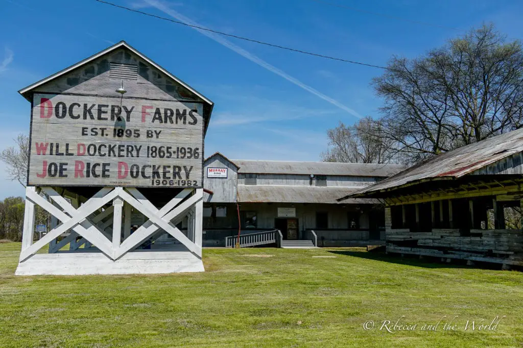 Exterior of Dockery Farms, established in 1895, with a large sign on a barn-like structure, surrounded by wide open grassy fields and clear skies. Dockery Farms is believed to be the birthplace of Delta blues music.