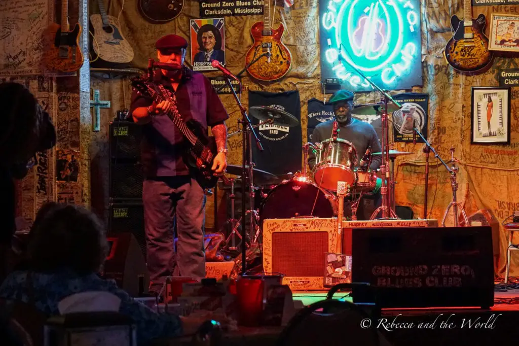 A dimly-lit blues club interior with a guitarist and drummer performing on stage, surrounded by walls covered in music memorabilia and neon signs. Clarksdale in Mississippi is the birthplace of the blues and you'll be able to catch live music most nights of the week. Live music at the Ground Zero Blues Club in Clarksdale, Mississippi.