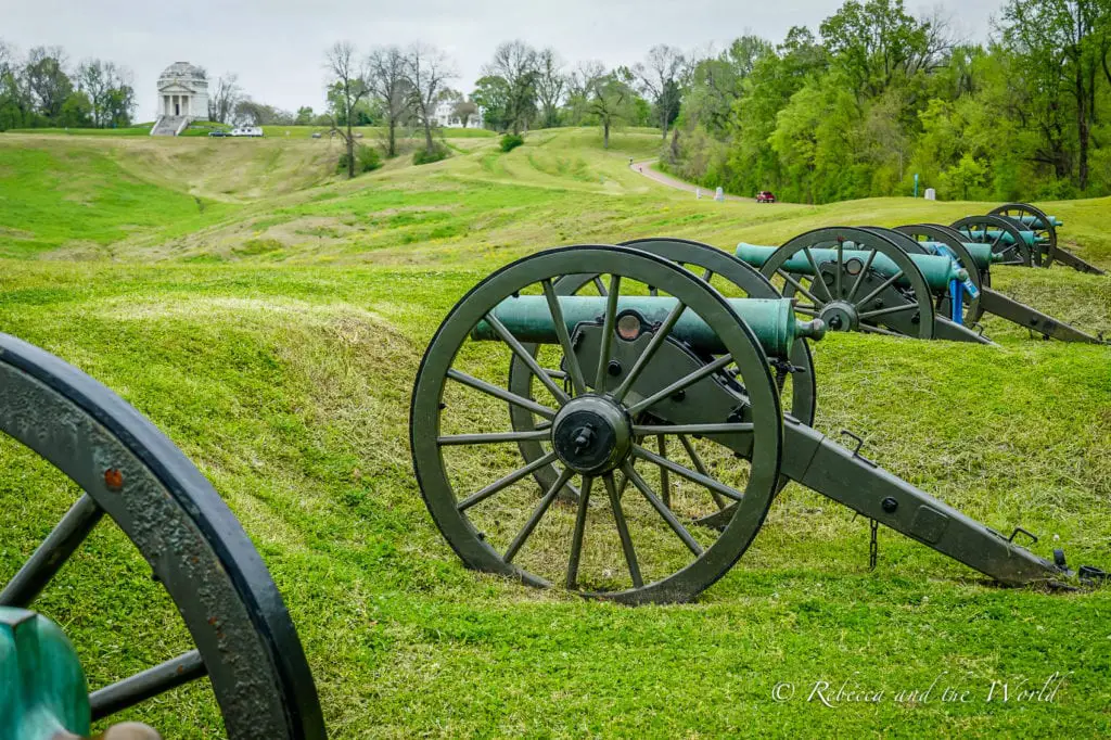 Row of historical cannons at Vicksburg National Military Park, with a monument and rolling hills in the background, under a cloudy sky. 