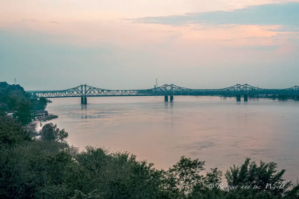 A serene view of a wide river with a long, multi-arched bridge spanning across. The bridge's reflection is visible in the calm water, and trees line the riverbank. The views over the Mississippi River in Natchez are stunning, especially at sunset. Sunset over the Mississippi River.